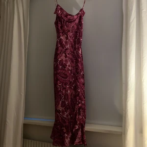 Mönstrad långklänning med volang - Säljer en långklänning med smala axelband och vackert paisleymönster i vinröda och rosa toner. Klänningen har volangdetalj nertill och en lätt, luftig känsla. Perfekt för sommarens tillfällen! Har inte använt den här någon gång och prislappen sitter kvar. 