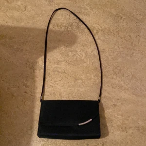 Svart axelväska med silverdetalj - Säljer en stilren svart axelväska med lång rem och elegant silverfärgad detalj på locket. Väskan har magnetlås och passar perfekt när du vill bära med dig det viktigaste. Enkel och tidlös design.