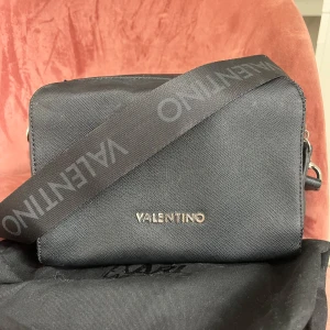 Svart från Valentino - Svart väska från Valentino med bred axelrem där märkesnamnet står tryckt. 