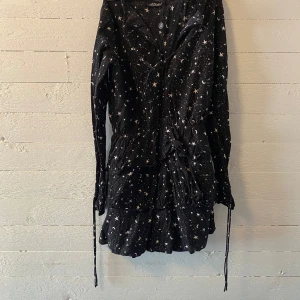 Svart klänning med stjärnmönster från MotelRocks - Svart klänning från MotelRocks med vitt stjärn- och mån-mönster.  Har långa ärmar, knytning i midjan och volangdetalj nertill. Aldrig använd! 🤗