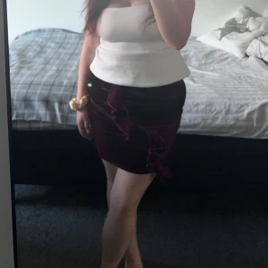 Vinröd sammetskjol med volang - Säljer en vinröd sammetskjol med snygg volangdetalj framtill. Kjolen är kort och har en figurnära passform som framhäver siluetten. 