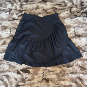 Svart omlottkjol med volang - Säljer en svart kort omlottkjol med volangdetalj. Kjolen har en snygg och trendig design med resår i midjan och lätt utsvängd form. 