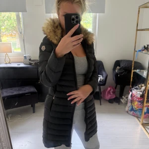 Moncler äkta päls  - Säljer en svart lång dunjacka moncler med äkta päls runt huvan. Jackan har dragkedja framtill och är quiltad för extra värme. Jätte fin men tyvärr blivit lite liten på mig