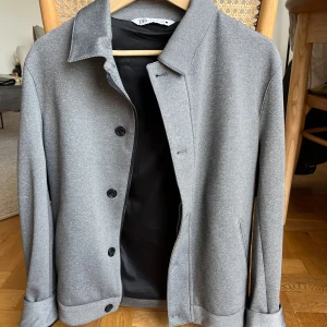 Grå jacka från Zara - Snygg grå jacka från Zara med klassisk krage och knäppning framtill med svarta knappar. Jackan har två sidfickor och är helfodrad. Perfekt för dig som gillar stilren och enkel design.