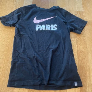 Svart Nike t-shirt med Paris-tryck - Svart t-shirt från Nike med stort rosa swoosh och texten PARIS på bröstet. Klassisk passform och korta ärmar. Perfekt för dig som gillar sportig stil och streetwear.