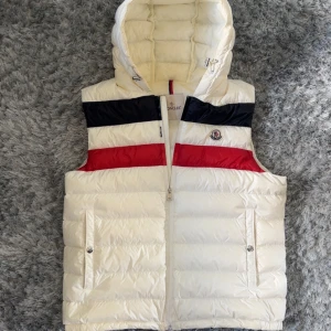 Moncler Timothe Gilet Väst herr vit - Snygg vit dunväst från Moncler med avtagbar huva och dragkedja framtill. Västen har svarta och röda detaljer över bröstet samt Moncler-logga på vänster sida. Två fickor med tryckknappar och justerbar dragsko i huvan. 1 flaw finns på baksidan, inget som stör men fick ej plats med ännu 1 bild, bild fås vid förfrågan!