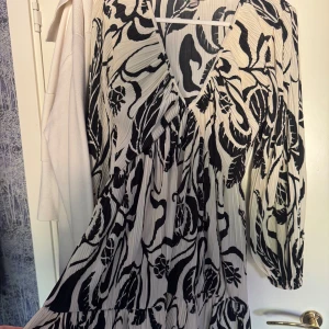 Svartvit plisserad klänning med v-ringning från h&m - Säljer en svartvit klänning med plisserat tyg och abstrakt mönster. Klänningen har v-ringning och långa ärmar med lös passform. Perfekt för dig som gillar unika prints och en luftig känsla.