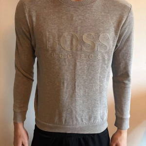 Grå sweatshirt från Hugo Boss - Säljer en stilren grå sweatshirt från Hugo Boss med diskret logga framtill. Tröjan har rund halsringning, långa ärmar och ribbade muddar vid ärmslut och nederkant. Perfekt för en avslappnad och snygg look.