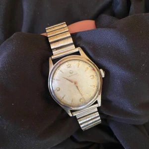 Certina herrklocka i silverfärg - Klassisk analog klocka från Certina med rund urtavla i vitt och silverfärgat armband i metall. Klockan har tunna visare. Perfekt accessoar för dig som gillar vintage stil.