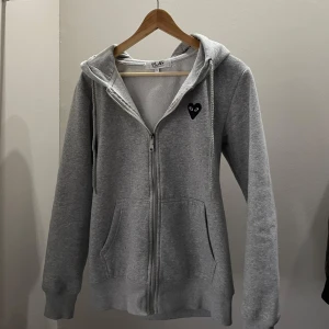 Grå hoodie med svart hjärta från Comme des Garçons Play - Grå hoodie med dragkedja och huva från Comme des Garçons Play. Tröjan har ett svart hjärta med ögon på bröstet och är tillverkad i mjukt material. Perfekt för en avslappnad stil. Priset är inte hugget i sten, skriv för fler bilder eller vid frågor 