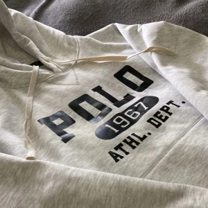 Grå hoodie från Polo Ralph Lauren - Säljer en ljusgrå hoodie från Polo Ralph Lauren med tryck 'POLO 1967 ATHL. DEPT.' på bröstet. Tröjan har huva med dragsko och ribbade muddar vid ärmslut och nederkant. Perfekt för en avslappnad stil.