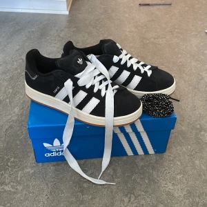 Svarta Adidas Campus sneakers - Säljer ett par svarta Adidas Campus sneakers med vita ränder och vit sula. Skorna har klassisk design med snörning och är i mocka. Medföljer extra svarta skosnören med vita detaljer. De är helt nya.