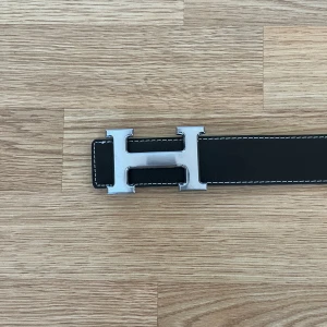 Svart bälte med silverfärgat H-spänne - Säljer ett svart bälte med ett stort, silverfärgat H-spänne. Bältet är gjort i skinn med vit kontrastsöm längs kanterna och har flera hål för justering. Perfekt accessoar för att lyfta din outfit. Helt ny och oanvänd men utan prislapp.
