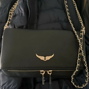 Svart axelväska med kjedja från Zadig & Voltaire - Snygg svart axelväska från Zadig & Voltaire med guldfärgade detaljer och kedja. Väskan har ett emblem med vingar framtill och dragkedja med märkets logga. Perfekt för att lyfta din outfit med en lyxig touch. Väskan är äkta och dustbagen kommer med. OBS inte mina bilder och väskan har en silvrig kjedja💞köparen står för frakten