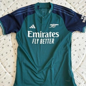 Arsenal fotbollströja från Adidas - Säljer en Arsenal fotbollströja från Adidas i grönt och mörkblått med vita detaljer. Tröjan har korta ärmar, klubbmärke och sponsortryck på bröstet. Perfekt för fotbollsfans! Tredjeställer 24/25 intill helt ny använd fåtal gånger. Storlek M