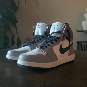 Nike Air Jordans i grått och vitt - Säljer ett par Nike Air Jordans med högt skaft i grått och vitt med svarta detaljer och svart swoosh. Skorna har klassisk snörning och rund tå. Perfekta för dig som gillar streetwear och vill sticka ut med en ikonisk sneaker vi kan prata om priset.