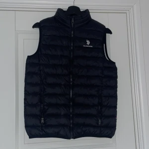 Svart dunväst från U.S. Polo Assn. - Säljer en svart dunväst från U.S. Polo Assn. Västen har hög krage, dragkedja framtill och två fickor med dragkedja. Broderad logga på bröstet. Perfekt för lager-på-lager och stilren look.