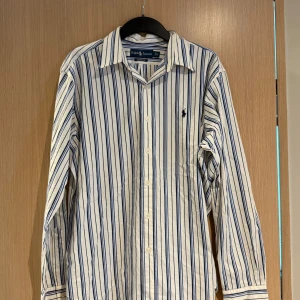 Randig skjorta från Ralph Lauren - Säljer en klassisk vit skjorta med blå ränder från Ralph Lauren. Skjortan har lång ärm, knappar framtill och den ikoniska broderade loggan på bröstet. Modellen är Classic Fit och passar perfekt till en stilren look.