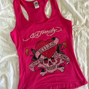 Ed Hardy Linne - Säljer ett äkta rosa linne från Ed Hardy med stort tryck framtill: dödskalle, hjärta och texten 'Love Kills Slowly' i glittriga detaljer. Brottarrygg och logga på ryggen. Perfekt & iconic statement-iconic Y2K linne 💓#y2k #mcbling #parishilton 