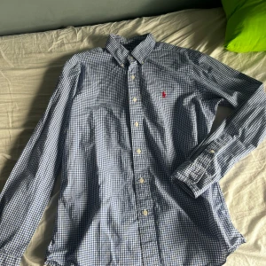Blårutig skjorta från Ralph Lauren - Säljer en klassisk blårutig skjorta från Ralph Lauren i modellen Custom Fit. Skjortan har lång ärm, knappar framtill och en röd broderad logga på bröstet. Perfekt för dig som gillar stilrena och tidlösa plagg.