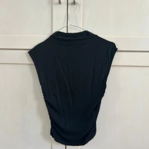 Svart topp från Gina Tricot - Säljer en svart topp från Gina Tricot i mjukt material. Toppen har korta ärmar och en snygg rynkad effekt på sidan som ger en smickrande passform. 