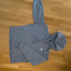 Grå hoodie från Acne Studios - Säljer en stilren grå hoodie från Acne Studios i storlek L. Tröjan är tillverkad i 100% bomull och har en klassisk huva med dragsko samt en liten broderad patch på bröstet. Perfekt för en avslappnad look. Pris kan diskuteras.