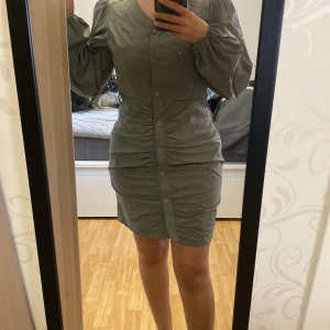 Grön kort klänning med puffärmar och knappar - Säljer en grön kort klänning med puffiga långa ärmar och knappar längs hela framsidan. Klänningen har en figurnära passform och lätt rynkade detaljer framtill. Perfekt för dig som gillar stilrena och trendiga plagg.