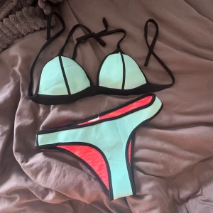 Triangl bikini  - Trendig bikini med trekantsöverdel och klassiska bikinitrosor. Framsidan är turkos med svarta kanter, medan baksidan är neonrosa. Knytband både upptill och nedtill för justerbar passform. Perfekt för stranden eller poolen!