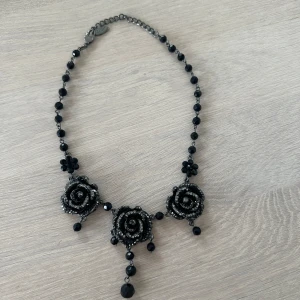 Svart halsband med rosor och pärlor - Unikt svart halsband med tre stora rosor dekorerade med svarta pärlor och glittriga detaljer. Halsbandet har små svarta pärlor längs kedjan och hängande pärlor under varje ros. Perfekt för att ge en dramatisk touch till din outfit.