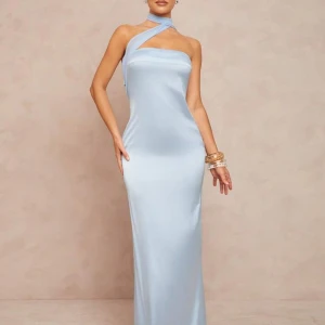 Ljusblå långklänning med asymmetrisk hals - Elegant ljusblå långklänning i satin med unik asymmetrisk halsringning och öppen rygg. Klänningen har en smal passform och ett långt band som knyts runt halsen för en modern look. Perfekt för speciella tillfällen.