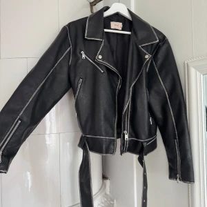 Svart skinnjacka från Nelly med bälte - Säljer en svart skinnjacka från Nelly i storlek M. Jackan har silvriga dragkedjor, markerade sömmar och ett bälte i midjan. Klassisk bikerstil med snedställd dragkedja och flera fickor.