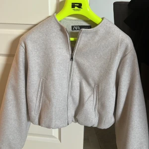 Ljusgrå croppad jacka från ZARA - Säljer en ljusgrå croppad jacka från ZARA med rund halsringning och dragkedja framtill. Jackan har långa ärmar och två fickor framtill. Endast använd 2 gånger så är i fint skick!!