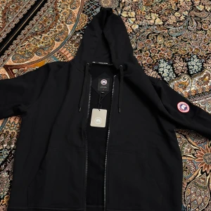 Svart hoodiejacka från Canada Goose - En sprillans ny hoodie från Canada Goose som är ganska varm och funkar perfekt för de lite kallare sommardagarna. Priset går att sänkas, vid snabb affär! Går även att lösa byxorna till Hoodien i samma storlek för endast 399!