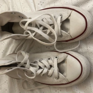 Vita höga Converse Chuck Taylor All Star - Klassiska vita höga Converse Chuck Taylor All Star med röd och blå rand längs sulan. Skorna har snörning, rund tå och platt sula. Perfekta för dig som gillar en tidlös och avslappnad stil.