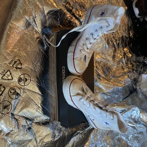 Vita höga Converse Chuck Taylor All Star - Klassiska vita höga Converse Chuck Taylor All Star med röd och blå rand längs sulan. Skorna har snörning och rund tå, tillverkade i canvas med Converse-logga på sidan. Perfekta för en avslappnad och tidlös stil.