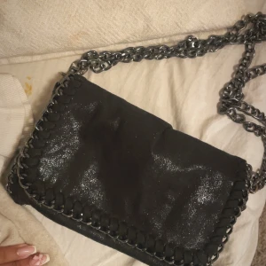 Svart axelväska med kedjerem - Säljer en svart axelväska med glittrig yta och dekorativa kedjedetaljer längs kanterna. Väskan har en rejäl kedjerem som ger en cool look och passar perfekt för att lyfta din outfit.