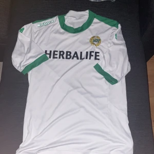 Hammarby T-shirt  - Säljer en vit Hammarby fotbollströja med gröna detaljer från Kappa. Tröjan har klubbmärke på bröstet och sponsortryck 'Herbalife' på framsidan. Klassisk design med gröna muddar och krage.
