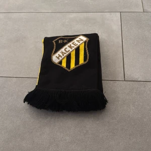 BK Häcken halsduk - Säljer en svart och gul halsduk från BK Häcken med klubbens logga och texten 'BK HÄCKEN' på båda sidor. Halsduken har fransar i ändarna och är perfekt för att visa ditt stöd på matchen.