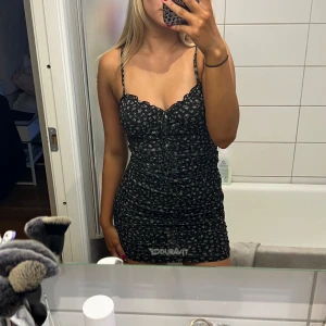 Svart blommig kortklänning med volangkant - Säljer en svart kortklänning med små vita blommor och volangkant upptill. Klänningen har smala axelband och en figurnära passform som framhäver midjan. 