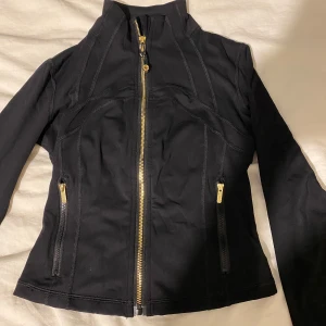 Svart jacka med dragkedja Lululemon  - Säljer en svart jacka med guldiga dragkedjor framtill och på fickorna. Jackan har en figurnära passform och hög krage. Perfekt för dig som vill ha en stilren och enkel jacka till vardags. Råkade ta fel storlek 