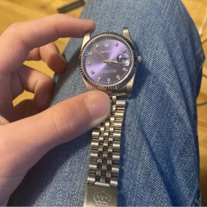 klocka - Säljer en snygg klocka från Pexor med lila urtavla och silverfärgad stållänk. Klockan har datumvisning och index med stenar. Klassisk rund boett och räfflad bezel. Passar dig som vill ha en stilren accessoar.