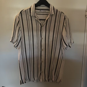 Vit och svart randig skjorta från Zara - Säljer en luftig vit skjorta från Zara med svarta vertikala ränder. Skjortan har korta ärmar, klassisk krage och knäppning framtill. Perfekt för varma dagar och enkel att styla med jeans eller shorts.