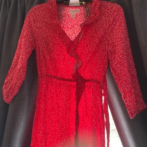 Röd omlottklänning med vita hjärtan från Holly & Whyte - Röd omlottklänning från Holly & Whyte med vita hjärtan, volangkant och trekvartsärm. Klänningen har knytband i midjan och är lätt och luftig, perfekt för varmare dagar.