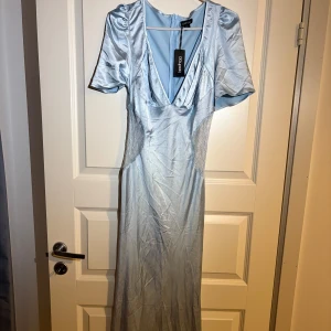 Balklänning - Säljer en elegant ljusblå långklänning från Boohoo med v-ringning och korta puffärmar. Klänningen har fina spetsdetaljer i sidorna och är tillverkad i ett glansigt satinliknande material. Perfekt för speciella tillfällen. Nyskick med prislappen på.