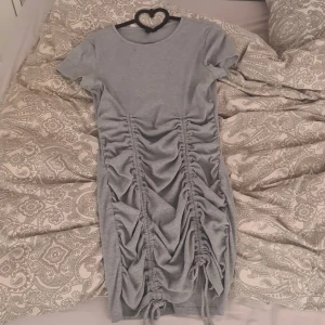 Grå ribbad klänning med rynkade detaljer från H&M Divided - Säljer en grå ribbad klänning från H&M Divided i storlek M. Klänningen har korta ärmar, rund halsringning och snygga rynkade detaljer med knyt framtill som ger en cool look. Perfekt för dig som gillar trendiga och bekväma plagg. Den är stretching och tjock samtidigt så kan man bestämma längden helt själv🎀