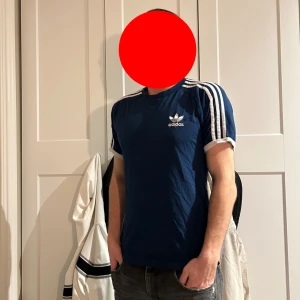 Blå Adidas t-shirt med vita ränder - Säljer en blå t-shirt från Adidas med klassiska vita ränder på axlarna och vitt Adidas-tryck på bröstet. T-shirten har korta ärmar och en sportig look, perfekt för dig som gillar streetwear.