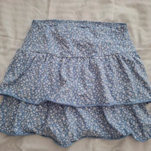 Blå blommig volangkjol från Lindex - Supersöt blå kjol med vitt blommönster från Lindex. Kjolen har volanger och är i en kort modell, perfekt för varma dagar. Tillverkad i ett lätt och skönt material som passar bra till sommaren.