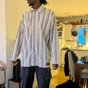 Randig skjorta från Karl Kani - Säljer en oversized skjorta från uncle sam med vertikala ränder i vitt, grått och brunt. Skjortan har långa ärmar, klassisk krage och knäppning framtill. Märkespatch nertill på framsidan. Perfekt för en avslappnad stil.