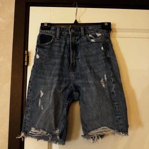 Blå jeansshorts med fransig kant - Säljer ett par blå jeansshorts med slitna detaljer och fransig kant nedtill. Klassisk femficksmodell med knappgylf och hög midja. Perfekta för en avslappnad stil.