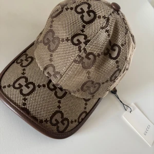 Beige och brun keps från Gucci - Säljer en beige och brun keps från Gucci med det klassiska GG-mönstret. Kepsen har böjd skärm med bruna detaljer och justerbar passform. Perfekt accessoar för att lyfta din stil.